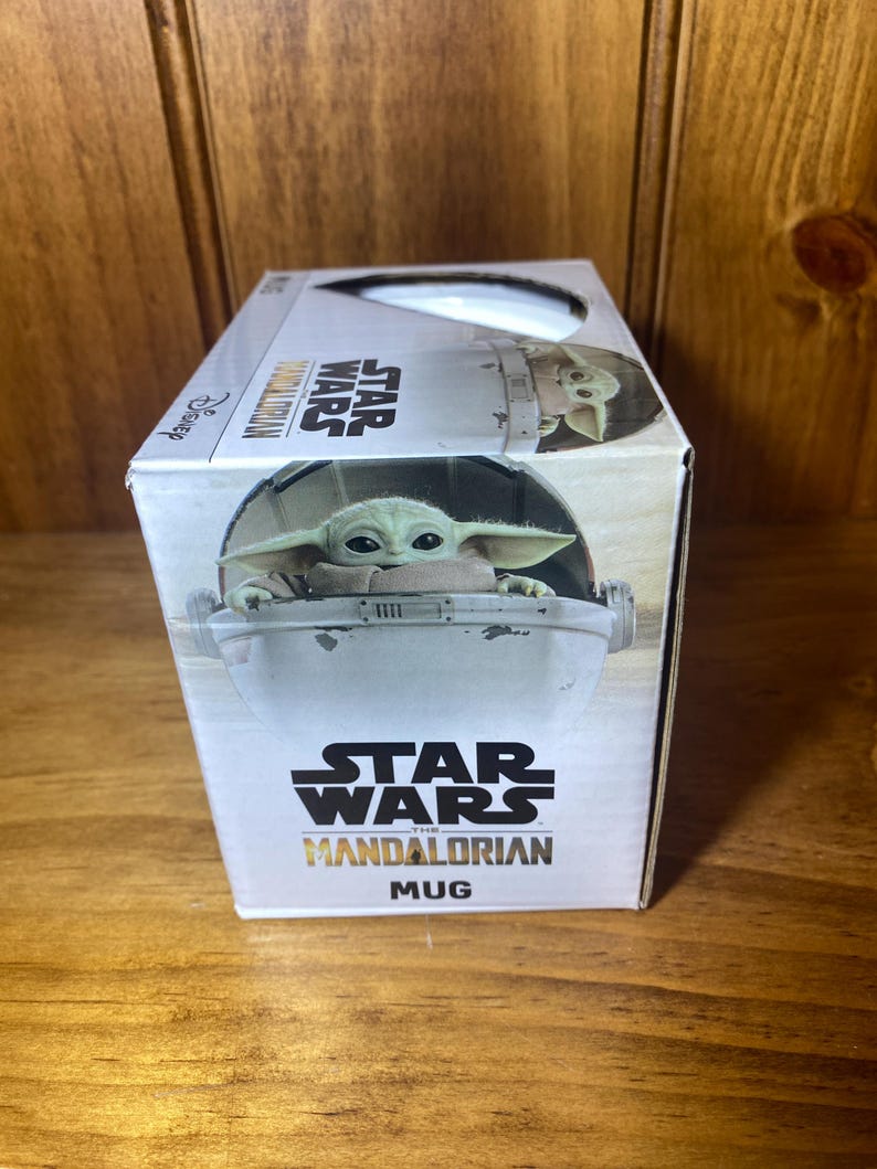 May include: A white box featuring a Star Wars Mandalorian mug. The box has an image of Grogu in his floating pram. The Star Wars logo and the word "Mug" are printed on the box.