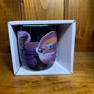 K&ouml;nnte beinhalten: Schwarze Keramik-Tasse mit einer Grafik von Grogu, auch bekannt als Baby Yoda, in seinem schwebenden Kinderwagen. Die Tasse befindet sich in einer wei&szlig;en Pappschachtel. Das Star Wars-Logo ist ebenfalls sichtbar.