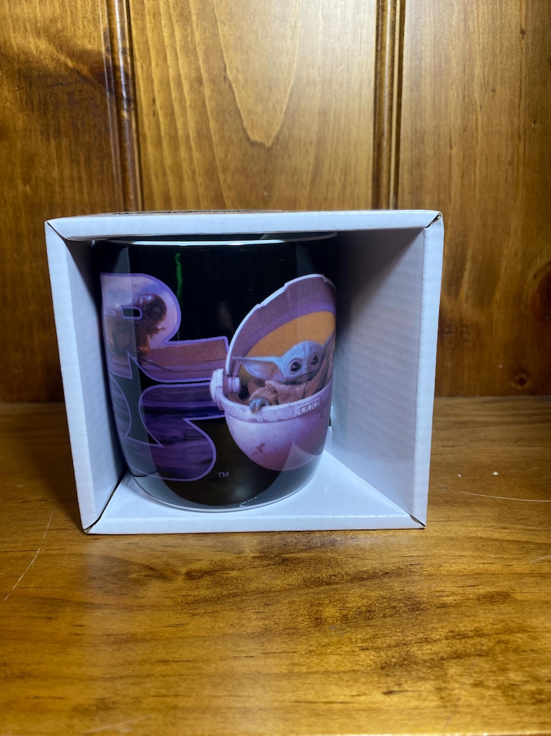 K&ouml;nnte beinhalten: Schwarze Keramik-Tasse mit einer Grafik von Grogu, auch bekannt als Baby Yoda, in seinem schwebenden Kinderwagen. Die Tasse befindet sich in einer wei&szlig;en Pappschachtel. Das Star Wars-Logo ist ebenfalls sichtbar.