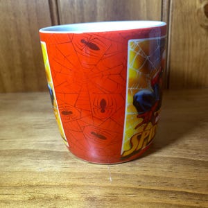 Puede incluir: Taza de cer&aacute;mica con Spider-Man. La taza tiene un fondo rojo con un patr&oacute;n de telara&ntilde;a y siluetas de ara&ntilde;as. El lado de la taza muestra a Spider-Man en acci&oacute;n. La taza es cil&iacute;ndrica con un interior blanco.