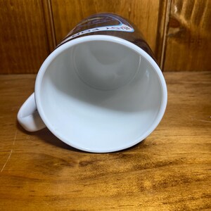 K&ouml;nnte beinhalten: Wei&szlig;e Keramik-Tasse mit Henkel, von oben betrachtet. Der Innenraum ist wei&szlig;, und die Au&szlig;enseite hat ein braunes Band mit Text und einem Design. Die Tasse steht auf einer Holzoberfl&auml;che.