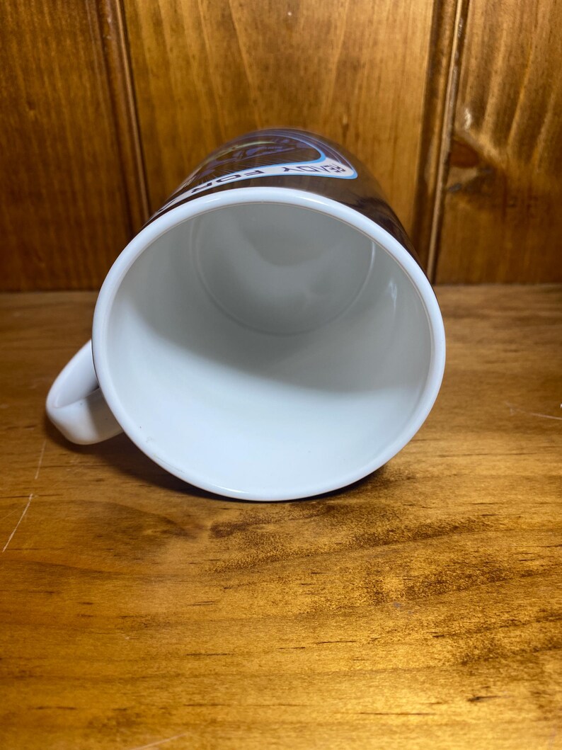 K&ouml;nnte beinhalten: Wei&szlig;e Keramik-Tasse mit Henkel, von oben betrachtet. Der Innenraum ist wei&szlig;, und die Au&szlig;enseite hat ein braunes Band mit Text und einem Design. Die Tasse steht auf einer Holzoberfl&auml;che.