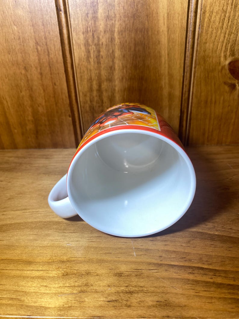 Puede incluir: Taza de caf&eacute; de cer&aacute;mica blanca con un dise&ntilde;o rojo y naranja. La taza tiene un asa blanca y un interior blanco. La taza est&aacute; sobre una superficie de madera.