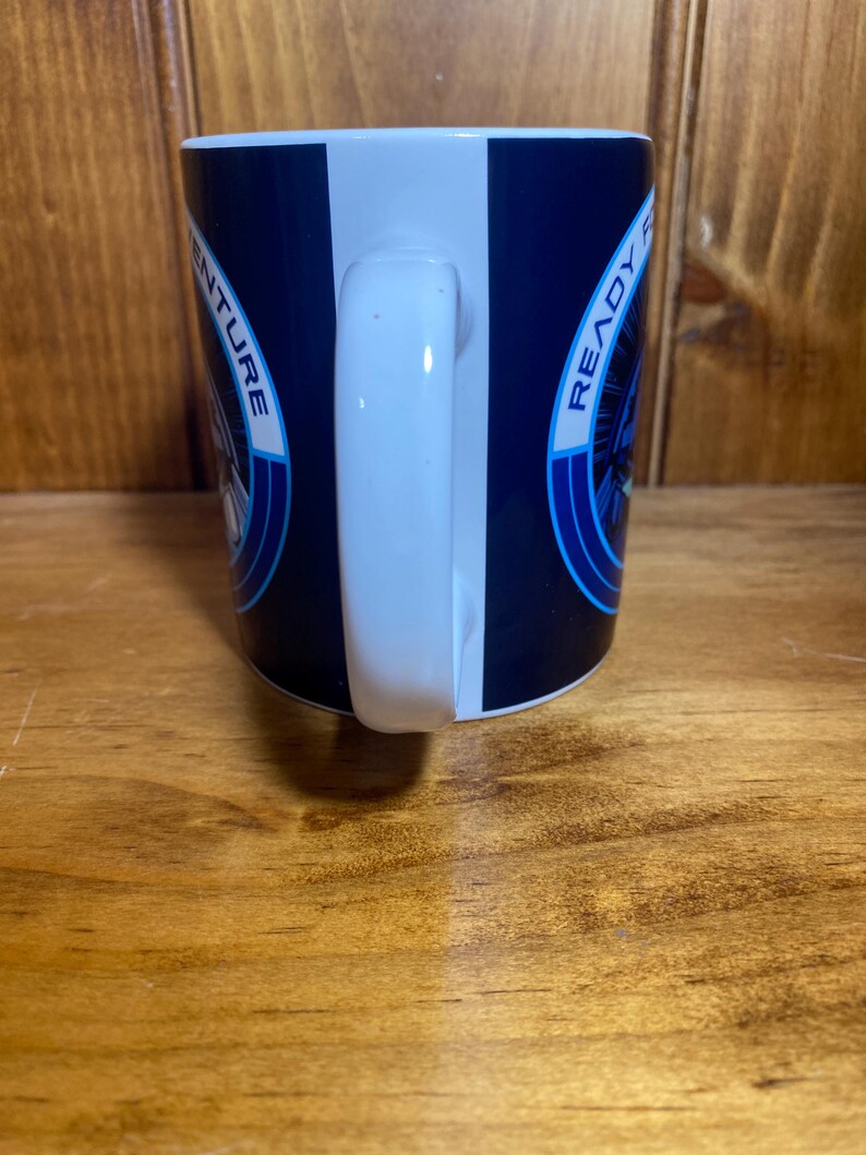 K&ouml;nnte beinhalten: Eine Kaffeetasse mit wei&szlig;em Griff, marineblauem Hintergrund und einem wei&szlig;en vertikalen Streifen. Die Tasse tr&auml;gt die Aufschrift "READY FOR ADVENTURE" und eine kreisf&ouml;rmige Grafik in Blau und Wei&szlig;.