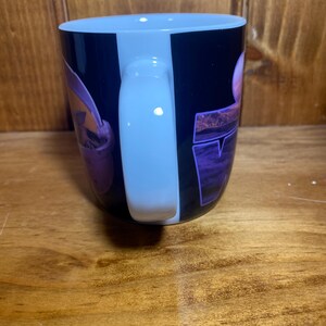 K&ouml;nnte beinhalten: Eine schwarze Kaffeetasse mit wei&szlig;em Henkel. Die Tasse hat ein lila und rosa Design mit dem Wort "TV" in lila Blockbuchstaben. Die Tasse steht auf einer Holzoberfl&auml;che.
