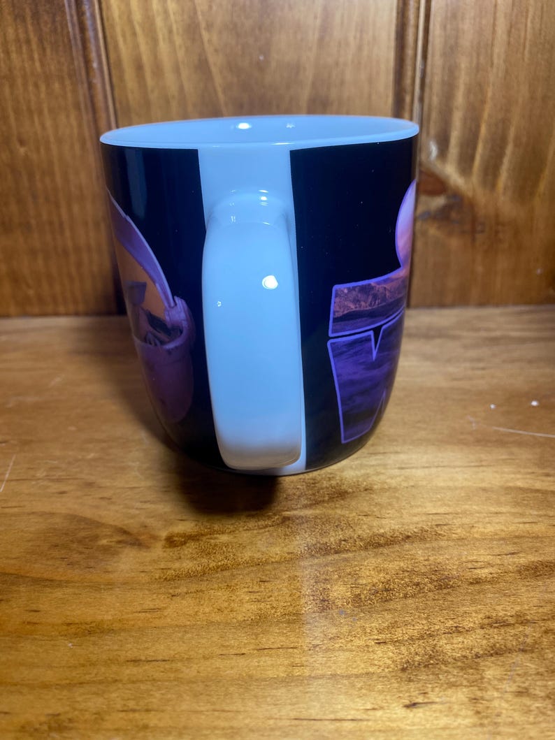 K&ouml;nnte beinhalten: Eine schwarze Kaffeetasse mit wei&szlig;em Henkel. Die Tasse hat ein lila und rosa Design mit dem Wort "TV" in lila Blockbuchstaben. Die Tasse steht auf einer Holzoberfl&auml;che.