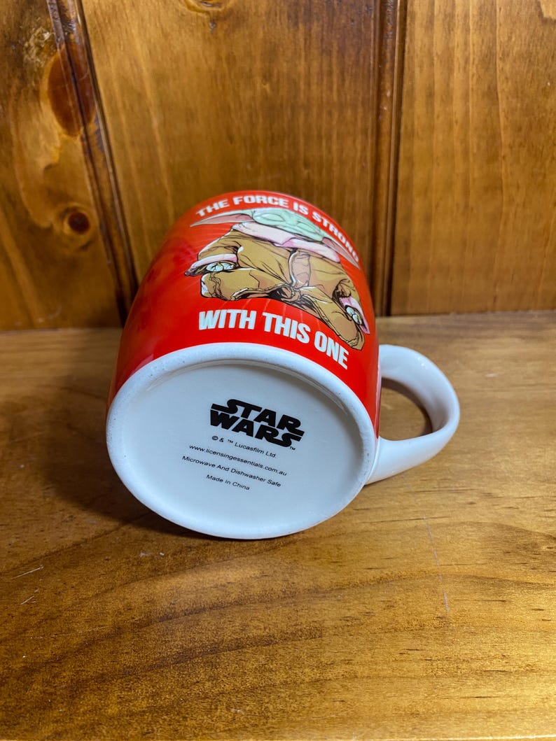 Pode incluir: Uma caneca de cer&acirc;mica vermelha com um gr&aacute;fico do Grogu e o texto THE FORCE IS STRONG WITH THIS ONE. A base exibe o log&oacute;tipo Star Wars, informa&ccedil;&otilde;es de direitos de autor e indica que a caneca pode ir ao micro-ondas e &agrave; m&aacute;quina de lavar.