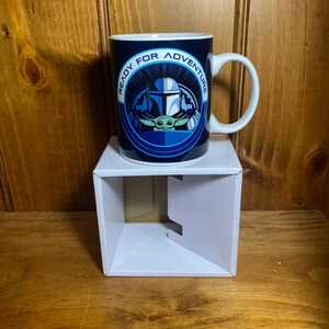 K&ouml;nnte beinhalten: Eine Keramik-Tasse mit blau-wei&szlig;em Design mit einem mandalorianischen Helm und Grogu, mit dem Text "READY FOR ADVENTURE". Die Tasse hat einen wei&szlig;en Henkel und steht auf einer wei&szlig;en Box.