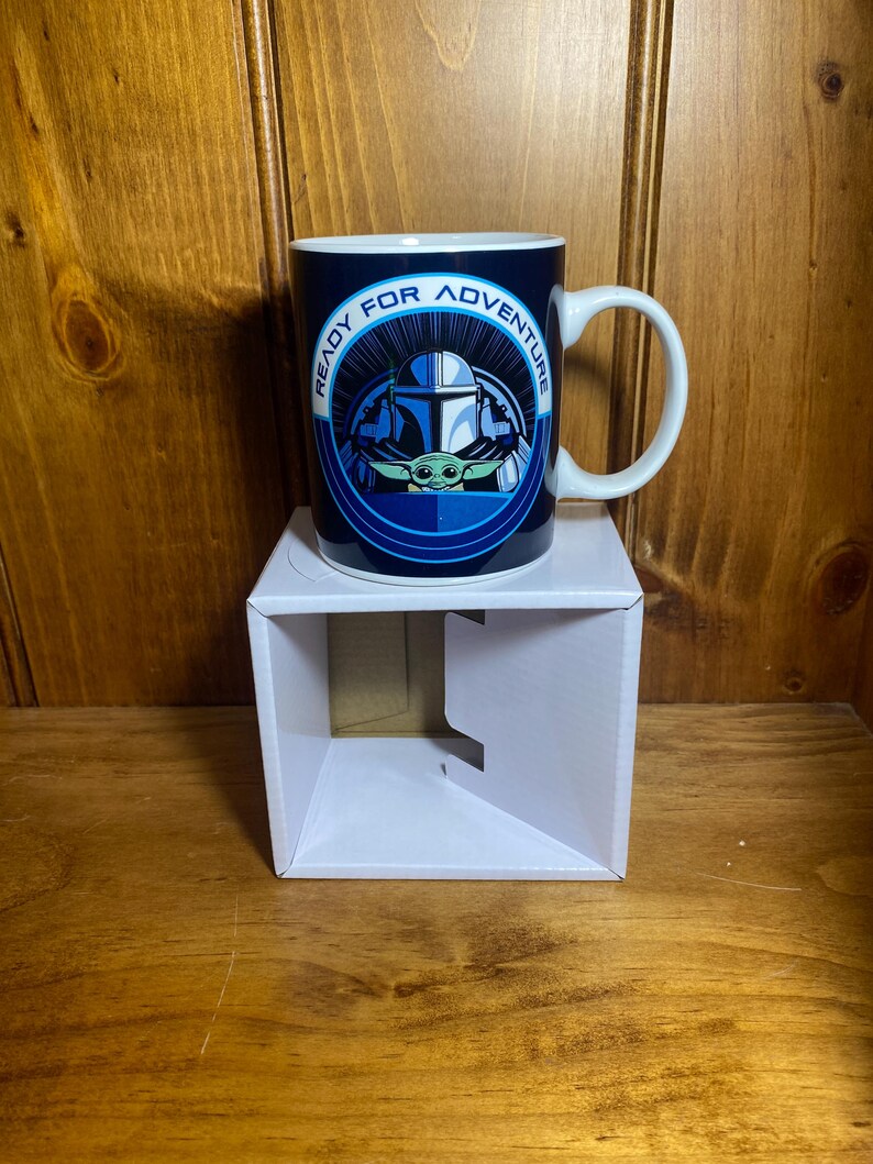 K&ouml;nnte beinhalten: Eine Keramik-Tasse mit blau-wei&szlig;em Design mit einem mandalorianischen Helm und Grogu, mit dem Text "READY FOR ADVENTURE". Die Tasse hat einen wei&szlig;en Henkel und steht auf einer wei&szlig;en Box.
