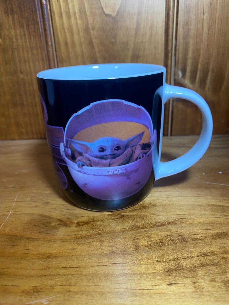 K&ouml;nnte beinhalten: Wei&szlig;er Keramikbecher mit einer Grafik von Grogu, auch bekannt als Baby Yoda, in seiner schwebenden Wiege. Der Becher hat einen schwarzen Hintergrund mit hellblauem Innenraum und Griff. Das Bild stammt aus der Star Wars-Serie The Mandalorian.