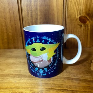 May include: A white ceramic mug with a blue background featuring a green alien character with large eyes. The mug has a white handle and the text "THIS IS THE WAY" in white lettering.