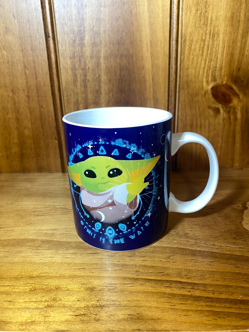 May include: A white ceramic mug with a blue background featuring a green alien character with large eyes. The mug has a white handle and the text "THIS IS THE WAY" in white lettering.