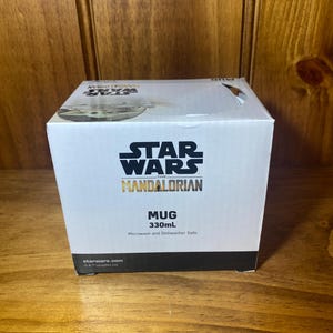 May include: A white box featuring the Star Wars: The Mandalorian logo. The box reads "MUG 330mL" and "Microwave and Dishwasher Safe." The box is sitting on a wooden surface.