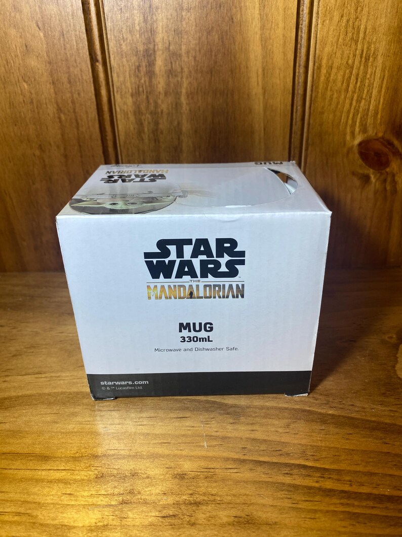 May include: A white box featuring the Star Wars: The Mandalorian logo. The box reads "MUG 330mL" and "Microwave and Dishwasher Safe." The box is sitting on a wooden surface.