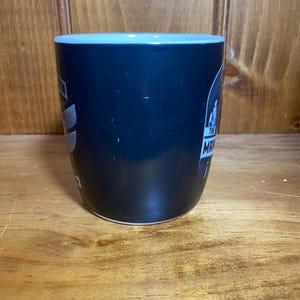 May include: A black coffee mug with a light blue rim and the text "The Man" in white. The mug has a glossy finish and is set against a wooden background.