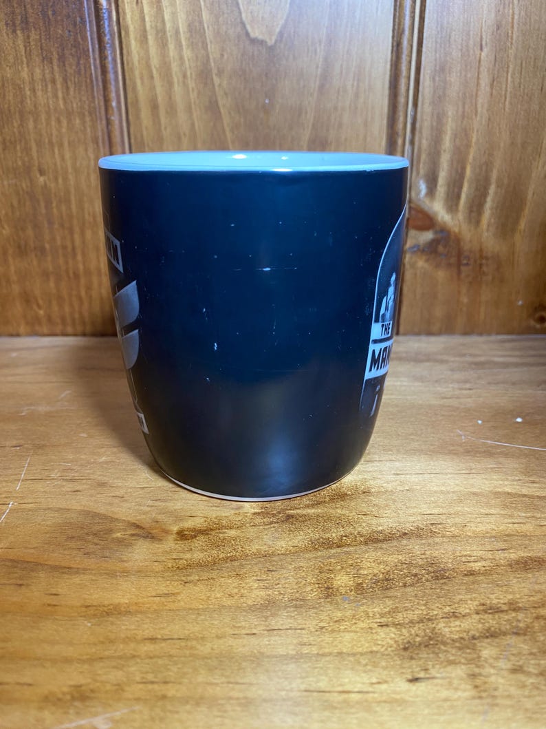 May include: A black coffee mug with a light blue rim and the text "The Man" in white. The mug has a glossy finish and is set against a wooden background.