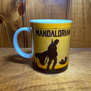 Puede incluir: Una taza de cer&aacute;mica amarilla con una silueta negra de The Mandalorian y Grogu en una c&aacute;psula flotante. El texto THE MANDALORIAN est&aacute; impreso en negro en la parte superior. La taza tiene un interior y un asa blancos, con un dise&ntilde;o de cielo estrellado.
