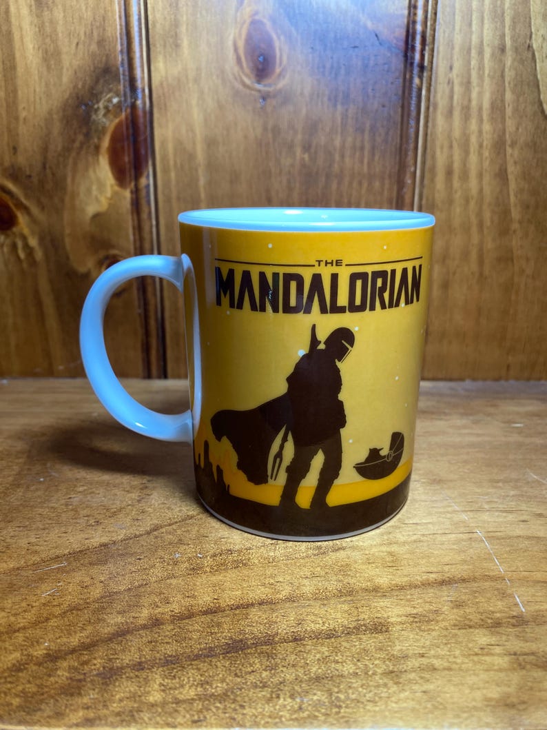 Puede incluir: Una taza de cer&aacute;mica amarilla con una silueta negra de The Mandalorian y Grogu en una c&aacute;psula flotante. El texto THE MANDALORIAN est&aacute; impreso en negro en la parte superior. La taza tiene un interior y un asa blancos, con un dise&ntilde;o de cielo estrellado.