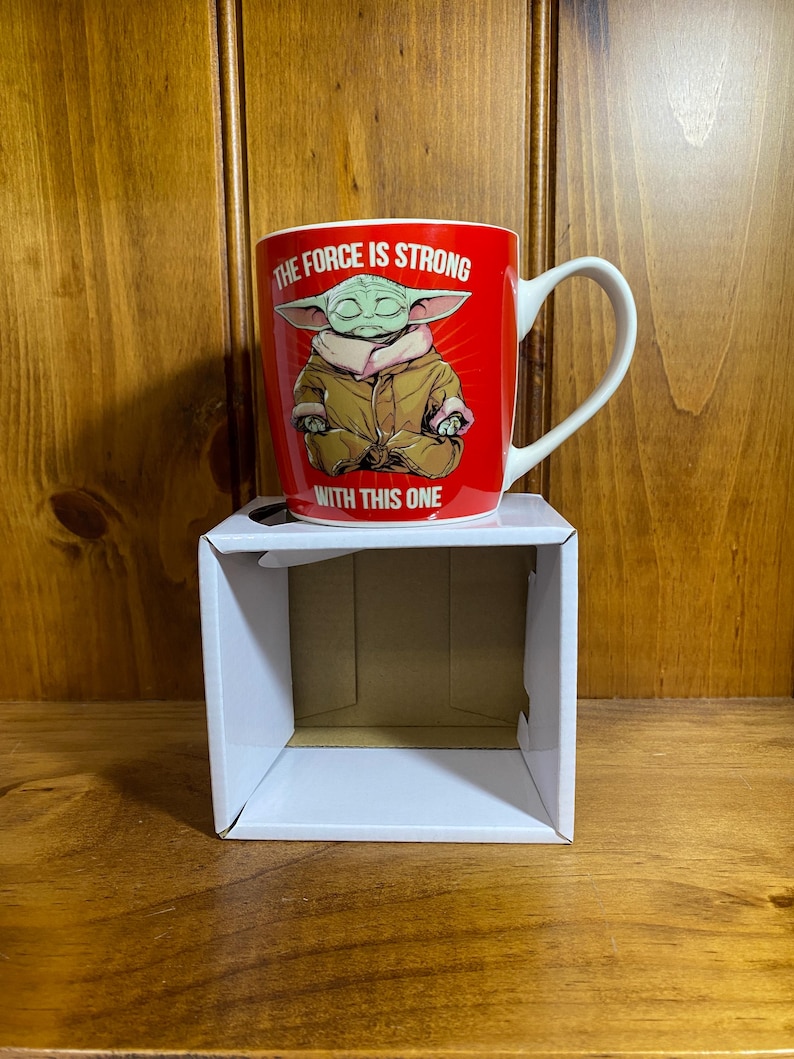 Pode incluir: Uma caneca de cer&acirc;mica vermelha com uma ilustra&ccedil;&atilde;o gr&aacute;fica de uma pequena criatura verde de orelhas grandes vestindo uma t&uacute;nica bege. O texto THE FORCE IS STRONG WITH THIS ONE est&aacute; impresso em letras mai&uacute;sculas brancas sobre a ilustra&ccedil;&atilde;o.