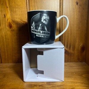 May include: Black ceramic mug featuring a graphic of The Mandalorian. The mug has a white handle and the text "THE MANDALORIAN THIS IS THE WAY" in white. The mug is sitting on a white cardboard box.