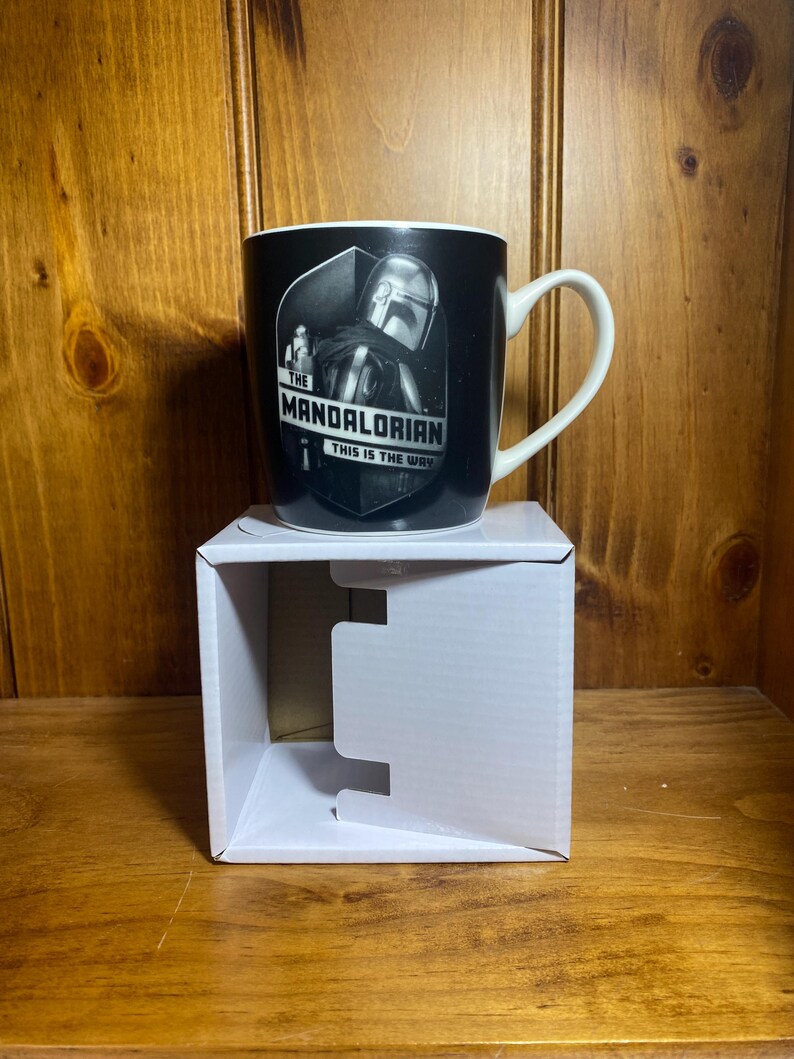 May include: Black ceramic mug featuring a graphic of The Mandalorian. The mug has a white handle and the text "THE MANDALORIAN THIS IS THE WAY" in white. The mug is sitting on a white cardboard box.