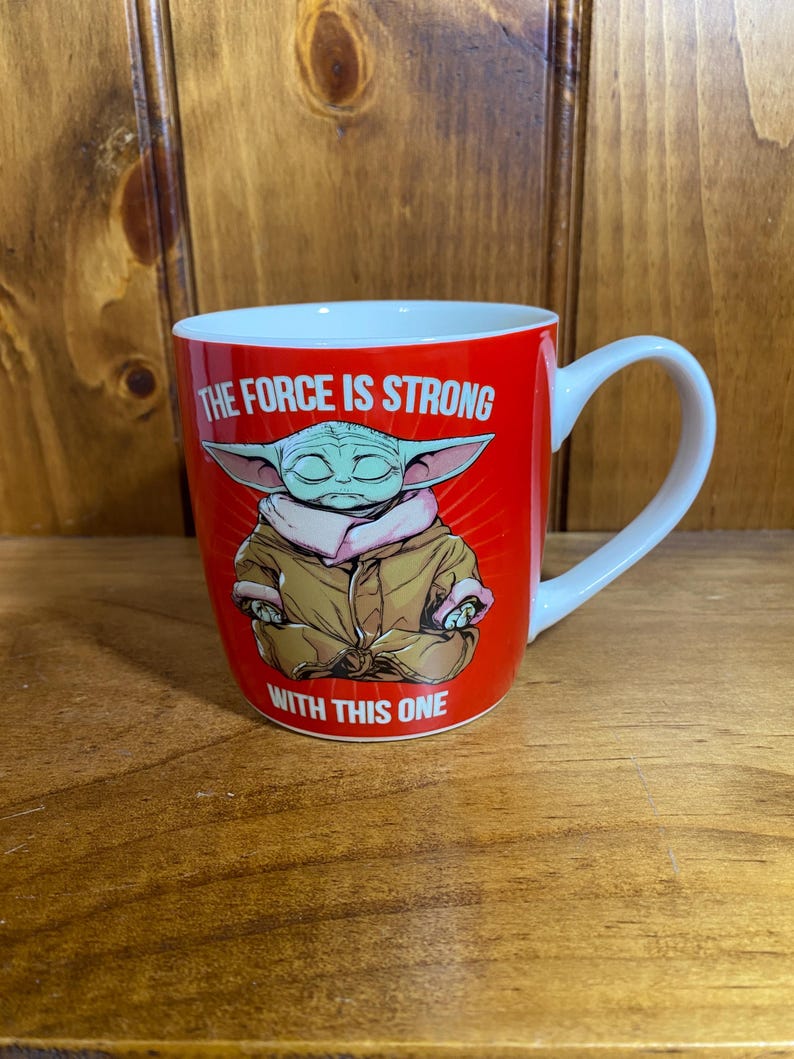 May include: A red ceramic mug featuring a graphic of a green, large-eared character in a tan robe sitting in a meditative pose. The text THE FORCE IS STRONG WITH THIS ONE is printed in white capital letters above and below the character illustration.