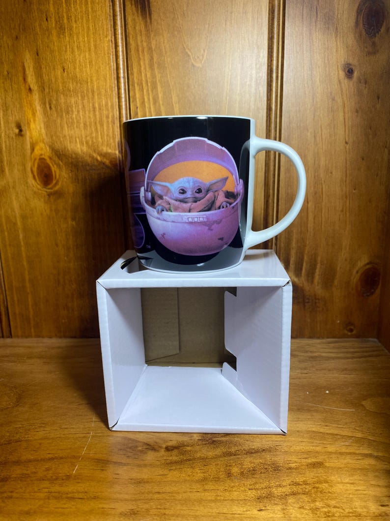 K&ouml;nnte beinhalten: Schwarze Keramik-Tasse mit einer Grafik von Grogu, auch bekannt als Baby Yoda, in einer rosa Trage. Die Tasse hat einen wei&szlig;en Henkel und steht auf einem wei&szlig;en Pappkarton. Der Hintergrund ist eine Holzoberfl&auml;che.