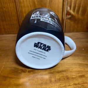 May include: Black ceramic mug featuring "The Mandalorian" and "This is the Way" text. The mug has a white handle and a white interior. The bottom of the mug displays the "Star Wars" logo and other text.