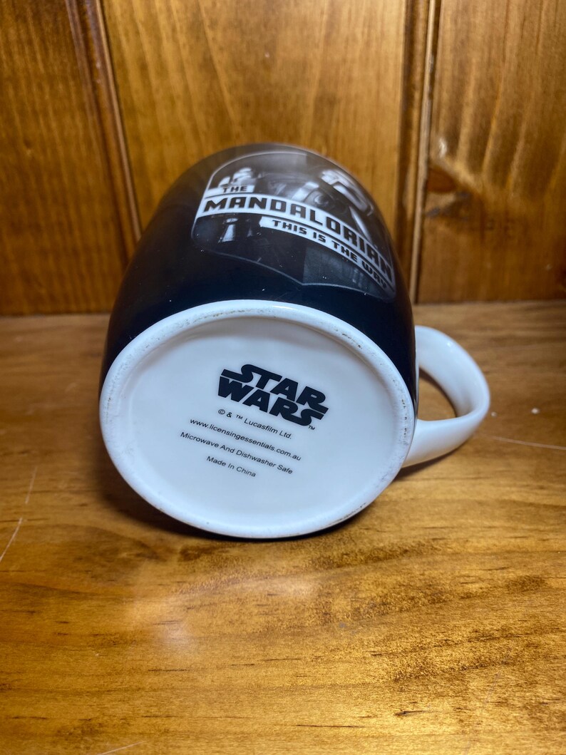 May include: Black ceramic mug featuring "The Mandalorian" and "This is the Way" text. The mug has a white handle and a white interior. The bottom of the mug displays the "Star Wars" logo and other text.