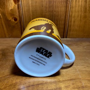Puede incluir: Una taza de cer&aacute;mica con un dise&ntilde;o en amarillo y marr&oacute;n que muestra la silueta de The Mandalorian. La base presenta el logotipo de Star Wars, texto que indica que es apta para microondas y lavavajillas, y Made in China.