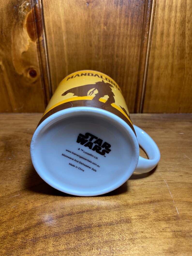 Puede incluir: Una taza de cer&aacute;mica con un dise&ntilde;o en amarillo y marr&oacute;n que muestra la silueta de The Mandalorian. La base presenta el logotipo de Star Wars, texto que indica que es apta para microondas y lavavajillas, y Made in China.