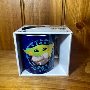 May include: A blue ceramic mug featuring a graphic of Grogu from The Mandalorian. The mug is in a white box with the word "MUG" printed on the side. The mug has a handle and is decorated with stars and the phrase "THIS IS THE WAY".
