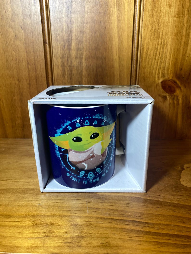 May include: A blue ceramic mug featuring a graphic of Grogu from The Mandalorian. The mug is in a white box with the word "MUG" printed on the side. The mug has a handle and is decorated with stars and the phrase "THIS IS THE WAY".
