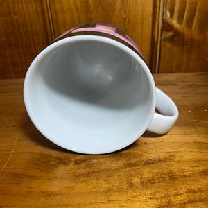 K&ouml;nnte beinhalten: Wei&szlig;e Keramik-Kaffeetasse mit Henkel. Die Oberseite der Tasse zeigt ein geometrisches Design in Braun-, Rosa- und Goldt&ouml;nen. Die Tasse steht auf einer Holzoberfl&auml;che.