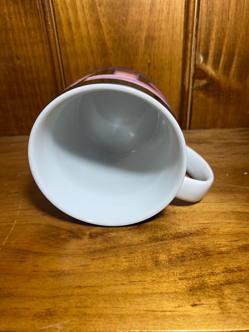 K&ouml;nnte beinhalten: Wei&szlig;e Keramik-Kaffeetasse mit Henkel. Die Oberseite der Tasse zeigt ein geometrisches Design in Braun-, Rosa- und Goldt&ouml;nen. Die Tasse steht auf einer Holzoberfl&auml;che.