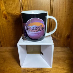 K&ouml;nnte beinhalten: Eine wei&szlig;e Keramiktasse mit einer Grafik einer Figur aus der Star Wars-Serie The Mandalorian. Die Figur ist in einem rosa-orangenen Tr&auml;ger vor schwarzem Hintergrund dargestellt. Die Tasse hat einen wei&szlig;en Henkel.