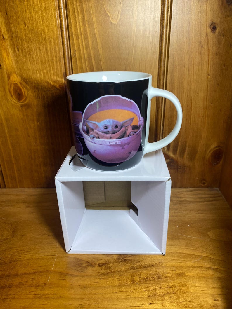 K&ouml;nnte beinhalten: Eine wei&szlig;e Keramiktasse mit einer Grafik einer Figur aus der Star Wars-Serie The Mandalorian. Die Figur ist in einem rosa-orangenen Tr&auml;ger vor schwarzem Hintergrund dargestellt. Die Tasse hat einen wei&szlig;en Henkel.