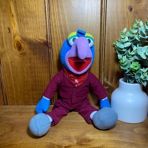Puede incluir: Un peluche del personaje Gonzo de Los Muppets, con cabeza azul, una nariz morada prominente, párpados amarillos y verdes, y una boca roja abierta. La figura viste una chaqueta y pantalones de color granate con una camisa roja debajo.