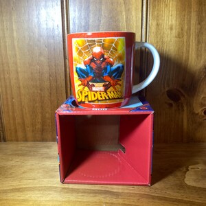 Puede incluir: Taza de cer&aacute;mica roja con Spider-Man en una pose din&aacute;mica, con el logo de Marvel. La taza tiene un asa blanca y se muestra en una caja roja con gr&aacute;ficos de Spider-Man. El fondo es una superficie de madera.