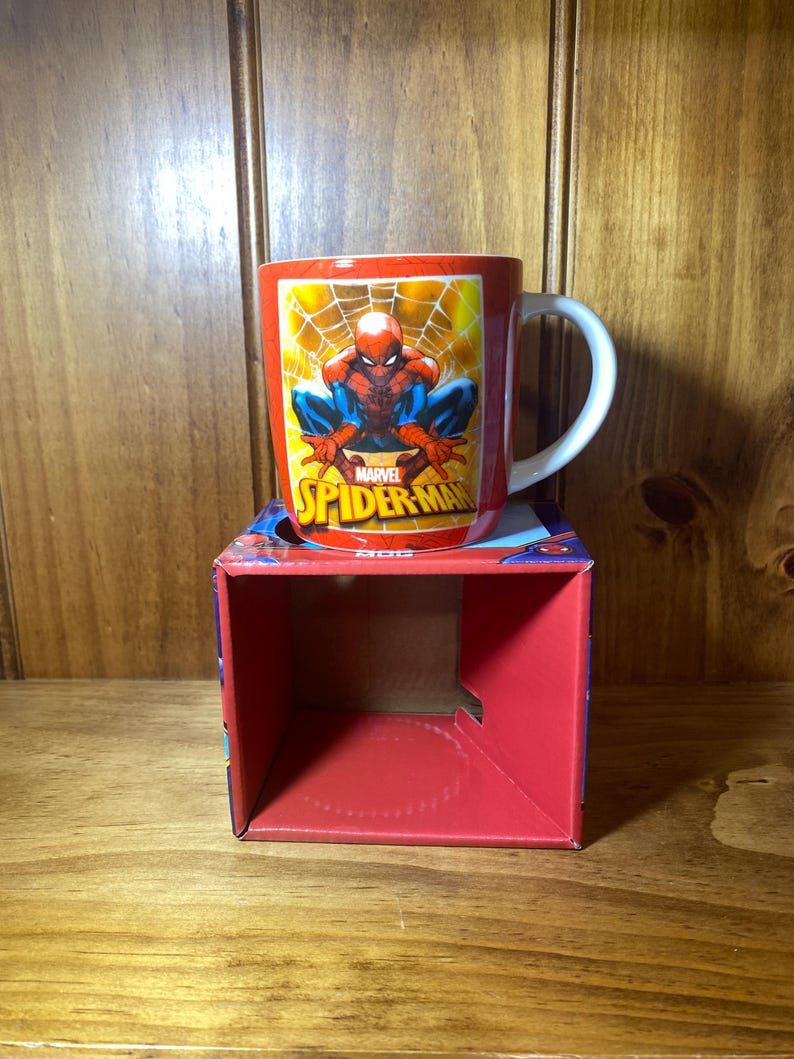 Puede incluir: Taza de cer&aacute;mica roja con Spider-Man en una pose din&aacute;mica, con el logo de Marvel. La taza tiene un asa blanca y se muestra en una caja roja con gr&aacute;ficos de Spider-Man. El fondo es una superficie de madera.