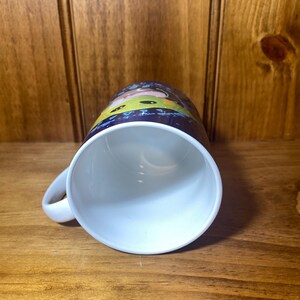 May include: White ceramic mug with a curved handle. The mug features a colorful cartoon design around the exterior. The interior of the mug is white. The mug is sitting on a wooden surface.