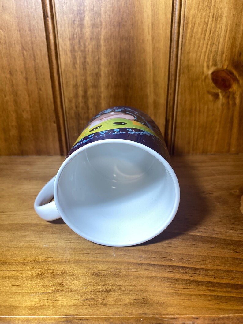 May include: White ceramic mug with a curved handle. The mug features a colorful cartoon design around the exterior. The interior of the mug is white. The mug is sitting on a wooden surface.