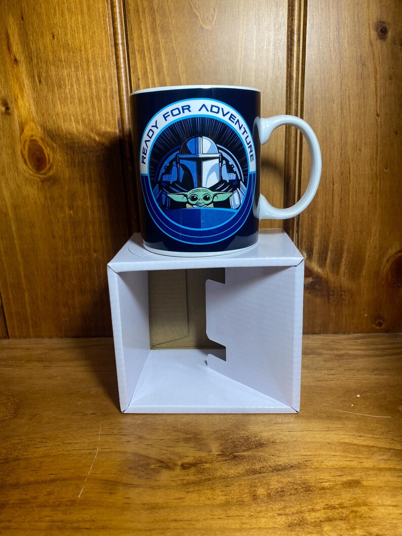 K&ouml;nnte beinhalten: Eine blaue Keramiktasse mit dem Text "READY FOR ADVENTURE", der ein Bild eines Mandalorianer-Helms und Grogu umgibt. Die Tasse hat einen wei&szlig;en Henkel und steht auf einem wei&szlig;en Karton.