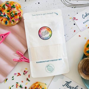 May include: A packet of Rainbow Party Biscuit Mix with a colourful logo and text. The image includes baked biscuits with rainbow sprinkles, pink clips, and a whisk. The text reads "Tear here to get started!"