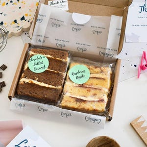 May include: A cardboard box containing two individually wrapped cake slices. One slice is chocolate with a green sticker labelled Chocolate & Salted Caramel, and the other is a yellow cake with a green sticker labelled Raspberry Ripple.