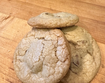 May include: Three golden brown sugar cookies on a wooden cutting board. Two of the cookies have a slightly raised center and the third cookie has a chocolate chip in the center.