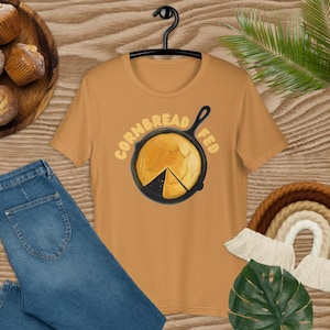 Puede incluir: Una camiseta amarilla mostaza con el texto "Cornbread Fed" en negro y una imagen de dibujos animados de una sartén de hierro fundido con una rebanada de pan de maíz dentro.