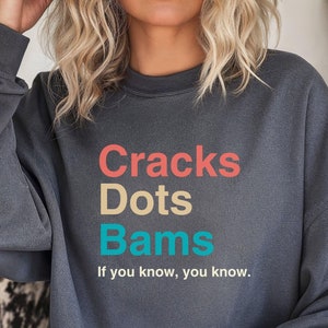 May include: A dark grey sweatshirt with the text "Cracks Dots Bams" in colourful letters. The text "If you know, you know." is printed below the main text.