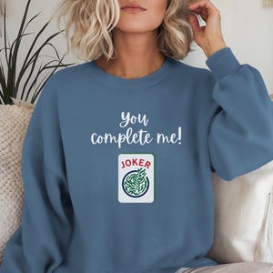 Puede incluir: Una sudadera azul con el texto "you complete me!" y un gr&aacute;fico de carta de joker verde y blanco.