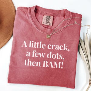 May include: A red-toned t-shirt with the text "A little crack, a few dots, then BAM!" in white. A silver necklace with a small pendant rests on the shirt. The shirt has a Comfort Colors tag.
